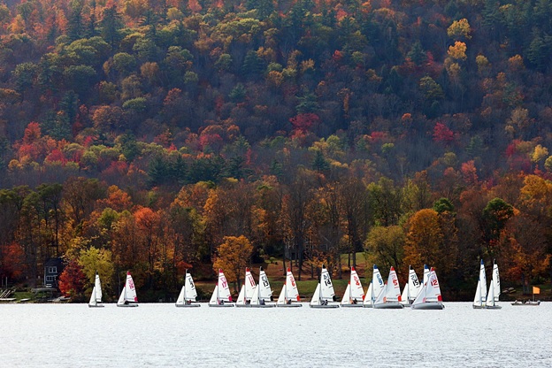 A group of small sailboats glides on a lake, surrounded by trees with vibrant autumn foliage in red, orange, and yellow hues, set against a forested hillside.