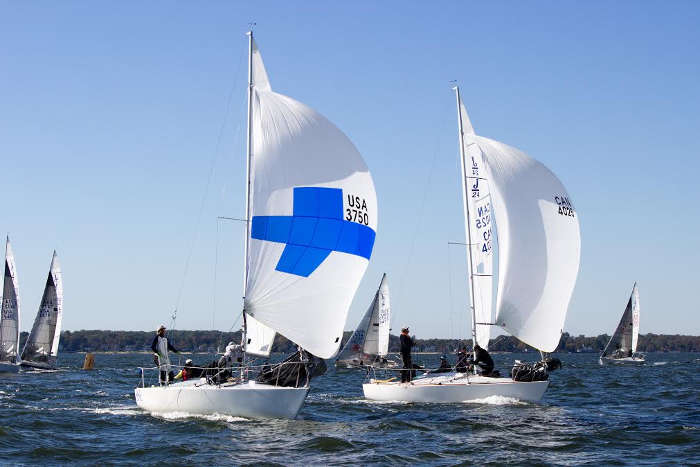 Two sailboats with white sails race on open water, each with crew members aboard. The left boat’s sail has a blue cross and 'USA 3750.' Other sailboats and a tree-lined shore are visible in the background.