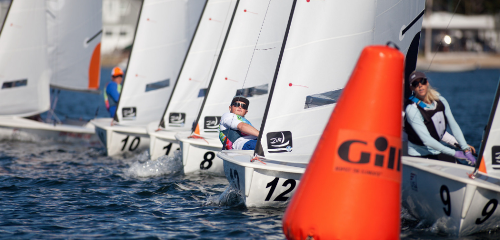 Several sailors compete in a race, maneuvering small sailboats around an orange buoy marked 'Gil' on open water, with their sails angled closely together. The sailors wear life vests and sunglasses.