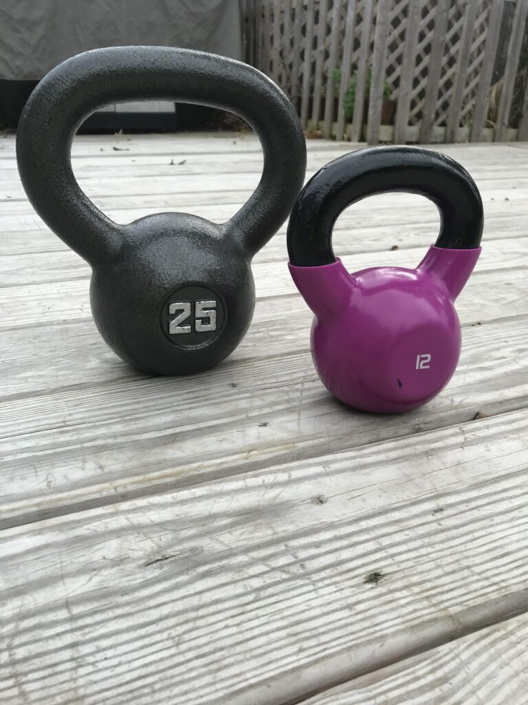 A 25-pound gray kettlebell and a 12-pound purple kettlebell are placed side by side on a wooden deck outdoors.