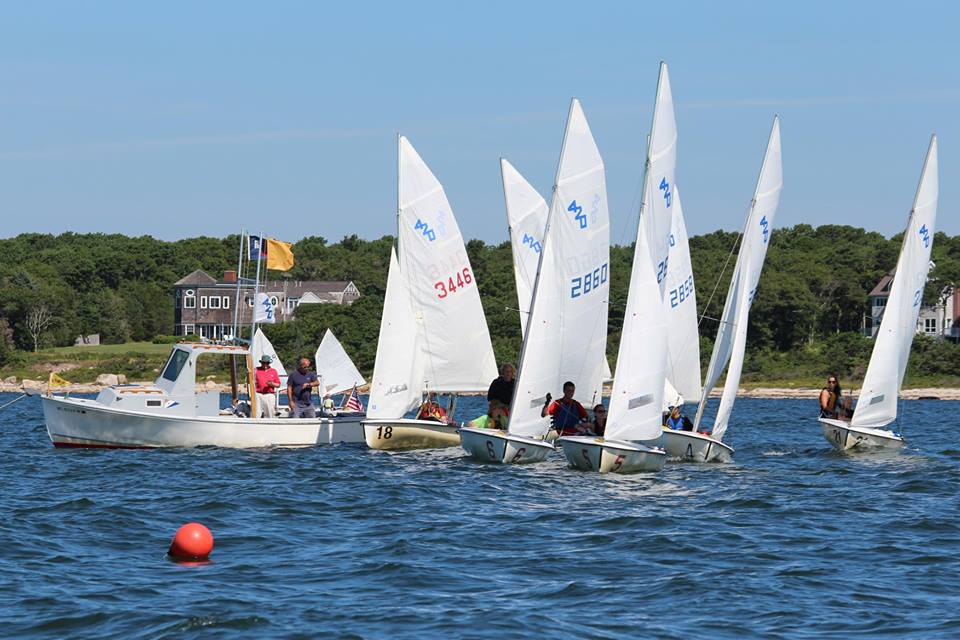Several small sailboats with white sails race near a motorboat with people aboard on a sunny day. The scene is set on open water with a shoreline, trees, and houses in the background. A red buoy floats in the foreground.