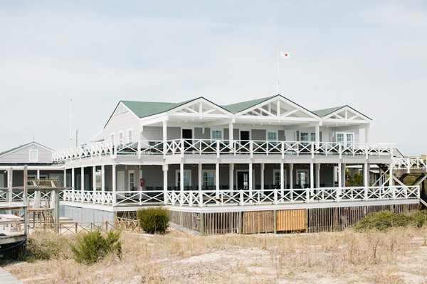 A large, white beach house with green roof, two stories, and wraparound porches featuring white railings, sits on sandy terrain with sparse grass under a cloudy sky.