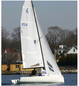 Two people sail a white J/70 sailboat on calm water near a shoreline with houses and trees visible in the background.