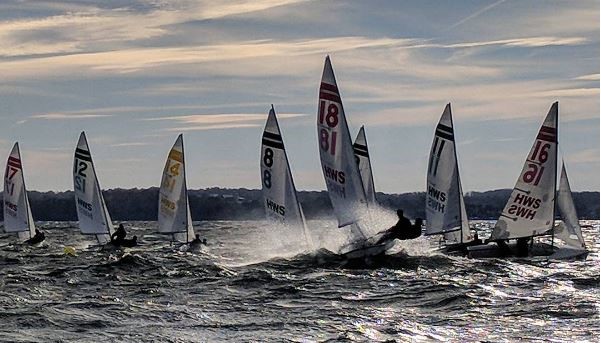 Several small sailboats with visible sail numbers race on choppy water under a partly cloudy sky. The boats tilt with the wind, and water sprays up as they move quickly. A distant shoreline is visible in the background.
