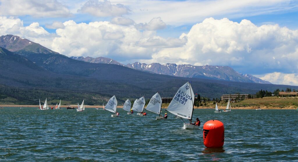 Small sailboats race on a lake surrounded by mountains under a partly cloudy sky. A large orange buoy floats in the foreground, while sailors navigate around it, and distant trees and buildings line the shore.