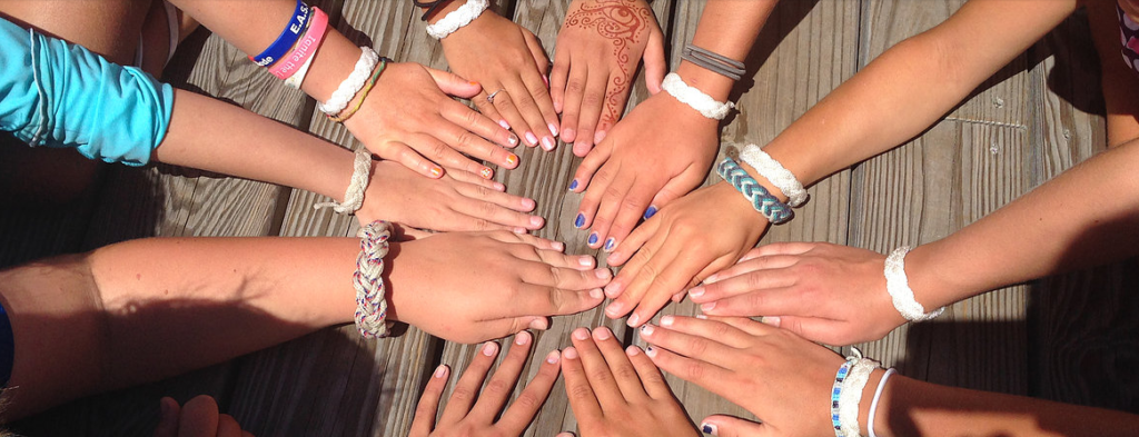 A group of people sitting in a circle place their hands together on a wooden surface, showing diverse skin tones, colorful nail polish, woven bracelets, and a henna design on one hand.