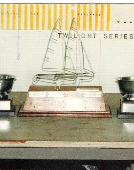 A metal wire sailboat trophy on a wooden base is displayed on a table between two silver cups. Behind them, a board reads "TWILIGHT SERIES" with a grid and yellow stripes at the top.