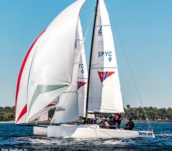 Two sailboats with white sails race on the water under a clear blue sky, with several people on board each boat. The sails show markings including "SPYC 9," and trees are visible in the background.