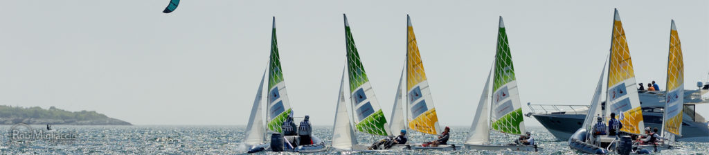 Several small sailboats with colorful sails and people aboard race on the water near a larger motorboat, while a kite surfer glides in the sky above on a sunny day. Distant shoreline is visible in the background.