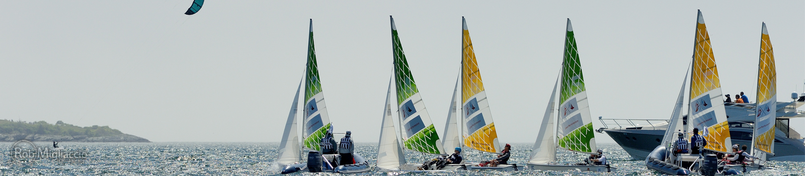 Several small sailboats with colorful sails and people aboard race on the water near a larger motorboat, while a kite surfer glides in the sky above on a sunny day. Distant shoreline is visible in the background.