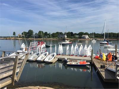 A small marina with several sailboats and motorboats docked at wooden piers on a calm, sunny day. White sailboats are lined up in a row, and houses and trees are visible across the water.