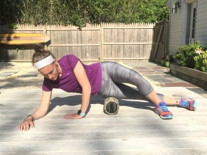 A woman in workout clothes uses a foam roller on her outer thigh while supporting herself on her side outdoors on a wooden deck.