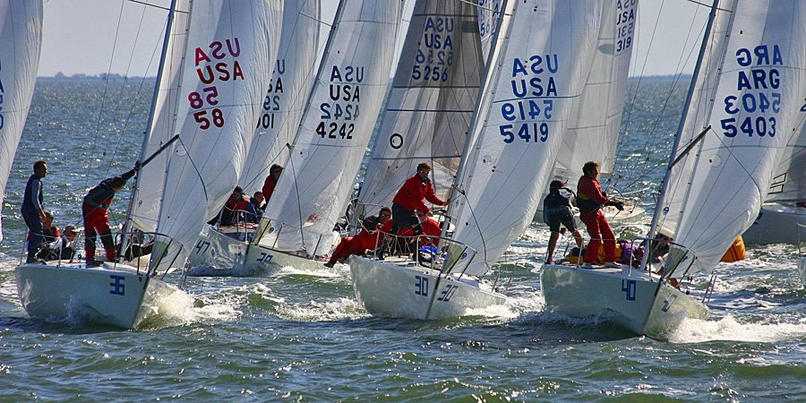 Several sailboats with crews racing closely on the water, sails marked with numbers and country codes, under a bright sky. The sailors are actively maneuvering as waves splash around the boats.