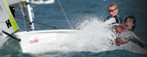 Two young sailors in life jackets on a small sailboat are splashed by a wave while navigating through choppy water, both appearing focused and exhilarated.