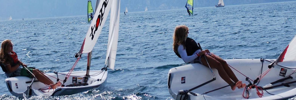Two people sail small boats on blue water, leaning back over the edge as they steer. The water is slightly choppy, and windsurfers and other sailboats are visible in the background. The scene is bright and outdoors.