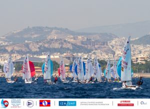 Sailboats with colorful sails compete in a regatta on the water, with a city and mountains visible in the background. Event sponsor logos appear along the bottom edge of the image.
