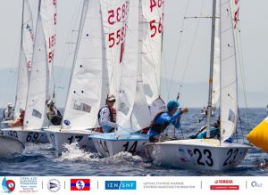A group of small sailboats with sailors compete in a regatta on open water. The boats are close together, and the sailors are focused. Event and sponsor logos appear along the bottom of the image.
