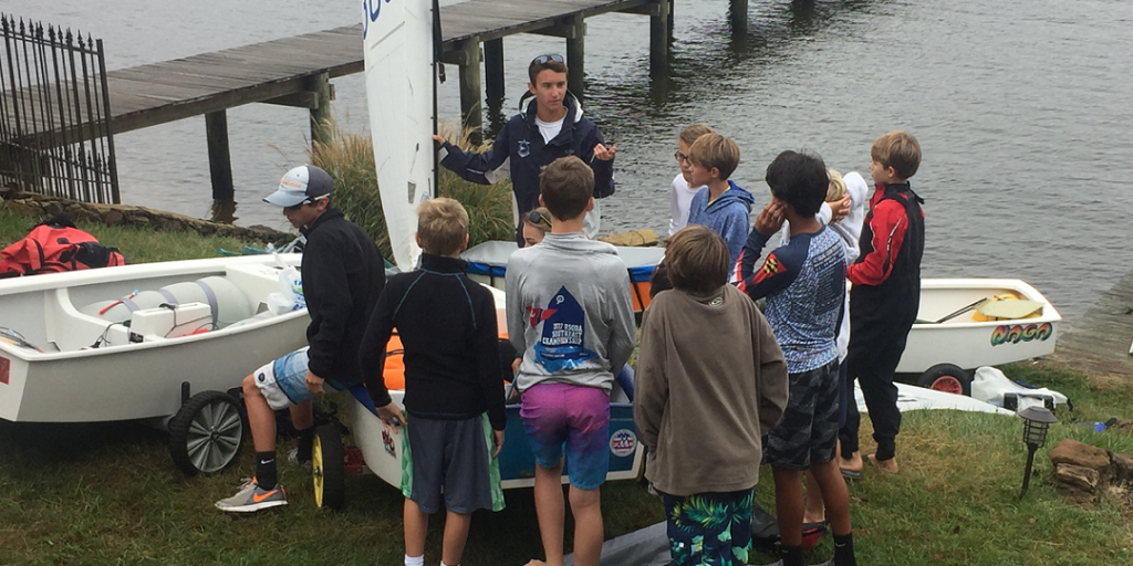 A group of children gather around an adult instructor by sailboats near a dock on a grassy shore, listening attentively as the instructor gestures and speaks.
