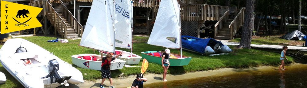 Children in life jackets stand by small sailboats on a grassy shoreline near a wooden dock and building; an ETYSA yellow flag with a black bear and crossed oars is visible on the left.