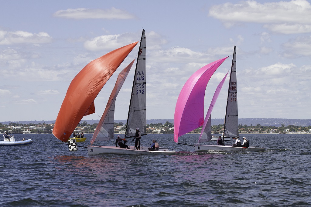 Two sailboats with colorful sails—one red and one pink—race side by side on a body of water, with people onboard each boat. A small motorboat and coastal buildings are visible in the background under a partly cloudy sky.