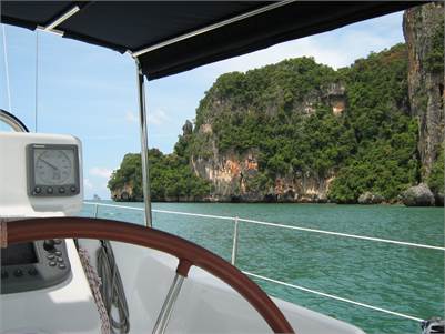 View from a sailboat showing a wooden steering wheel, navigation instruments, and metal railing with turquoise water and lush green cliffs in the background under a partly cloudy sky.