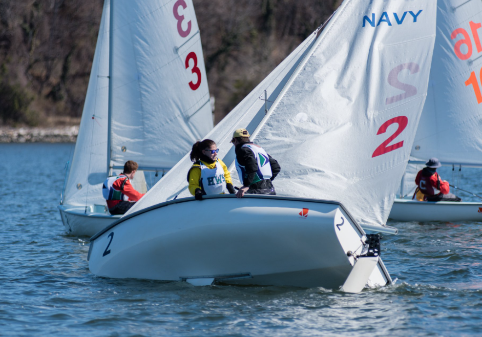Two sailboats with visible "NAVY" and number "2" on their sails compete on a sunny day. Four sailors are aboard, wearing life jackets and gear, navigating close to each other on calm water. Trees are in the background.
