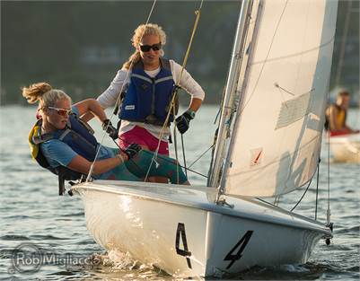 Two people wearing life vests sail a small boat marked with the number 4, leaning out to balance as they navigate the water on a sunny day. Another boat is visible in the background.