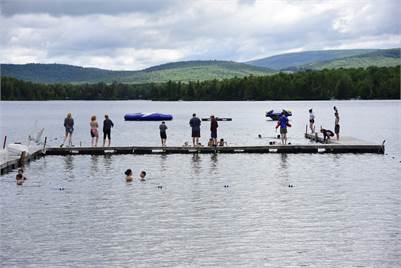 People stand, swim, and play on and around a dock extending into a lake, surrounded by forested hills under a cloudy sky. Some are jumping into the water; others watch or relax on the dock.