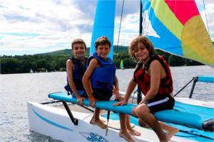 Three smiling children wearing life jackets sit on the edge of a small sailboat on a lake, with colorful sails, water, and trees in the background.