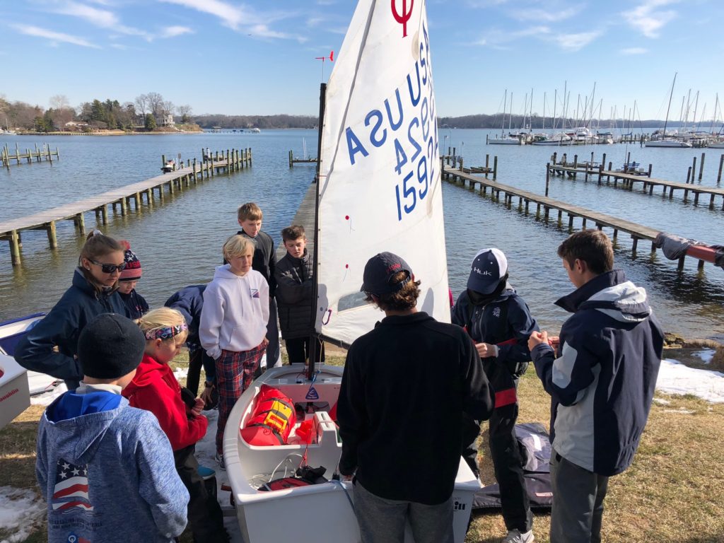 A group of kids and a few adults gather around a small sailboat near a dock on a lake, preparing for sailing on a sunny day with clear skies and many boats in the background.