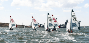 Several small sailboats with numbers on their sails race on choppy water under a partly cloudy sky, with industrial structures visible in the background.