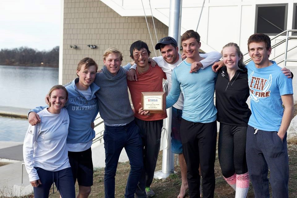 Seven smiling young adults stand together outdoors by a lake, some with arms around each other. One person in the center holds a plaque. They appear to be celebrating and are dressed casually.