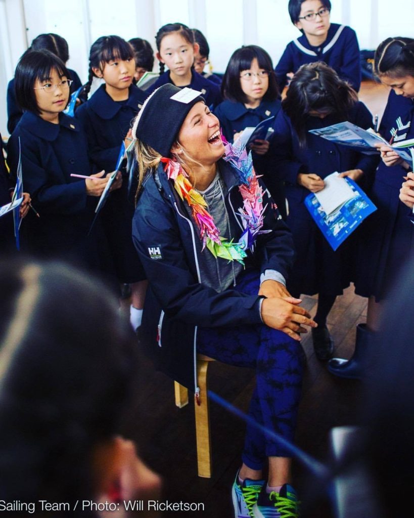 A smiling woman wearing a colorful paper crane necklace sits on a chair, surrounded by a group of schoolchildren in uniforms, who are holding papers and looking at her with interest.