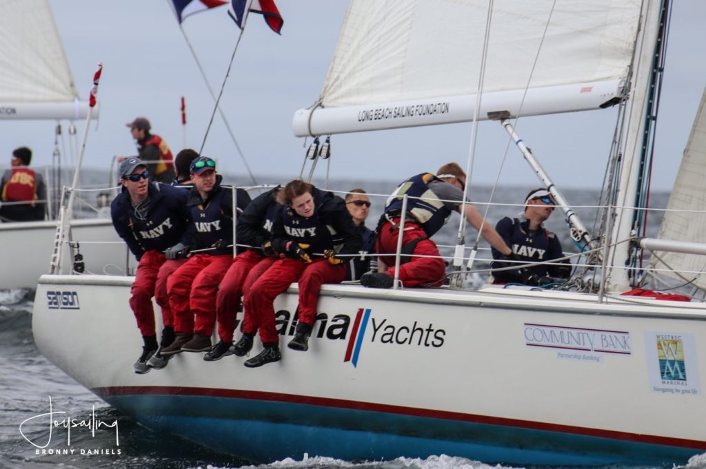 A group of young sailors in red pants and navy shirts, labeled 'NAVY,' sit on the edge of a white sailboat racing on the water. The boat displays sponsor logos and flags fly in the background.