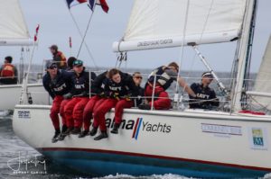 A group of young sailors in red pants and navy shirts, labeled "NAVY," sit on the edge of a white sailboat racing on the water. The boat displays sponsor logos and flags fly in the background.