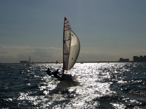 A small sailboat with two people aboard sails on a sunlit sea, with sparkling water and a bright sky. Buildings and other boats are visible on the distant horizon.