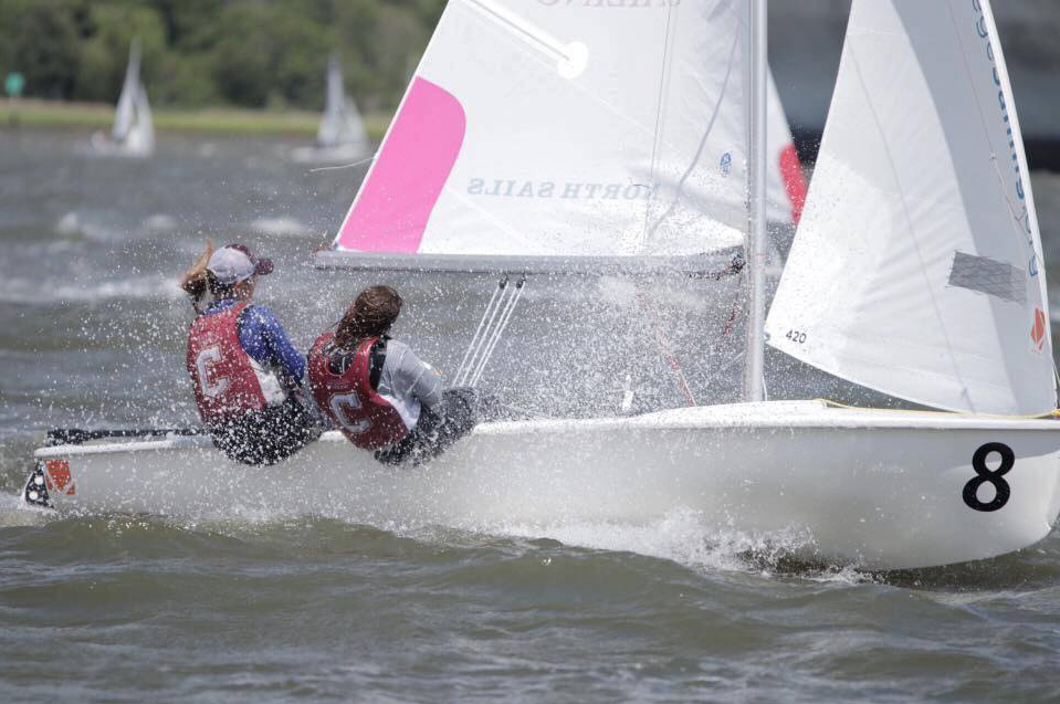 Two sailors wearing life vests and numbered jerseys steer a small white sailboat numbered 8 through choppy water, with a spray of water splashing up and other sailboats visible in the background.
