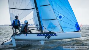 Two people sailing a small catamaran with blue and white sails on open water; the boat number 15 and the word 'NACRA' are visible on the hull. Both sailors are wearing helmets and life vests.