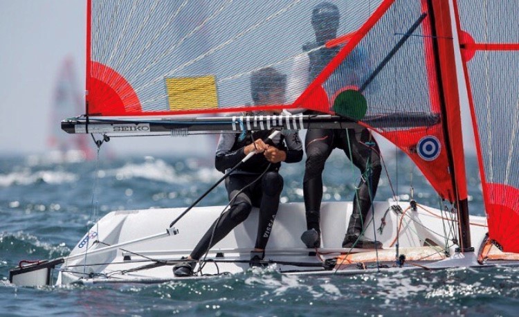 Two sailors wearing wetsuits maneuver a small sailboat with red sails across choppy water during a race. The ropes and rigging are visible, and another sailboat is seen in the distance.