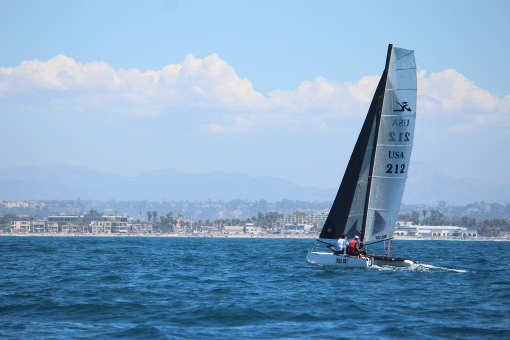 A small sailboat with two people sails on blue water near a coastal city, with buildings, palm trees, and mountains visible in the background under a partly cloudy sky.