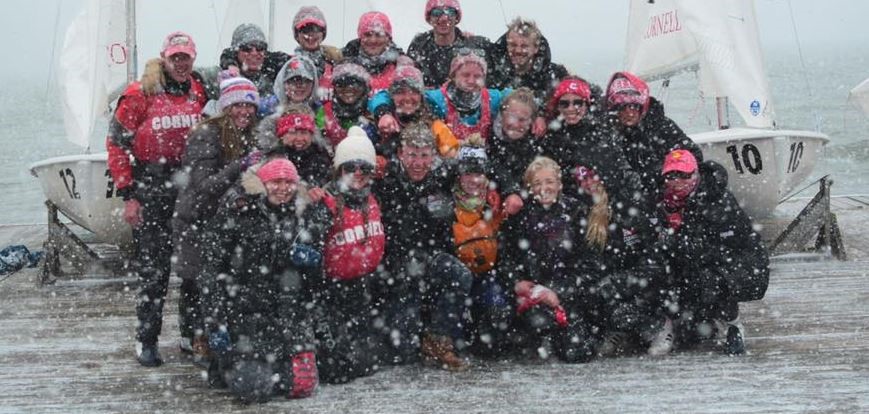 A large group of people in winter clothing pose together on a dock in falling snow, with sailboats behind them.