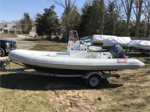 A small inflatable boat with an outboard motor sits on a trailer parked on grass, with trees and other boats in the background.
