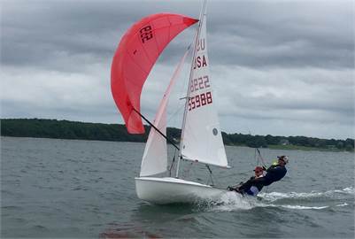 Two people sail a small white dinghy with a red spinnaker on a cloudy day. They are leaning out over the water to balance the boat, with a forested shoreline visible in the background.