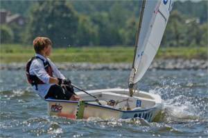 A young person wearing a life jacket sails a small white dinghy on a river or lake, with water splashing at the bow and green trees visible in the blurred background.