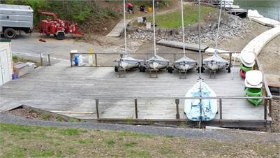 Five small sailboats are docked upright on a wooden platform near a shoreline. A blue kayak and several green paddle boats are also on the dock. A curved path and rocky slope lead down to the water. Trees surround the area.