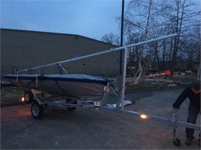 A small sailboat on a trailer with its mast partially raised is being moved by a person in an outdoor area near a warehouse at dusk. Trees and various items are visible in the background.