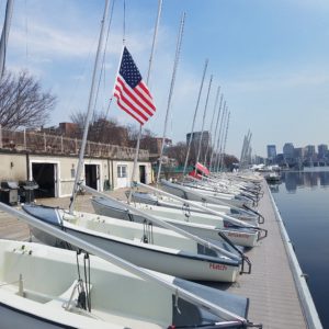 A row of small sailboats docked along a pier, with an American flag flying overhead. Buildings and trees are visible in the background across the calm water under a blue sky.