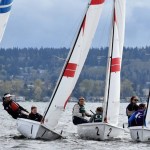 Several sailboats with teams of people are racing on a lake, leaning to balance against the wind. The background shows trees, houses, and a cloudy sky. The boats are marked with numbers.