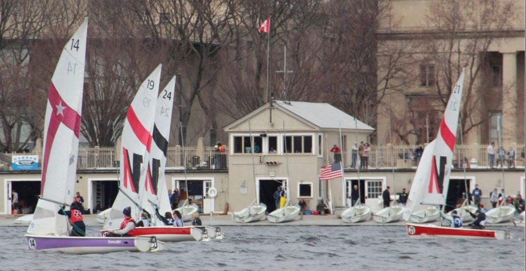 Several small sailboats with 'N' and '14' or '19' on their sails race on a choppy river. People watch from the dock and a waterside building, with leafless trees and large buildings in the background.