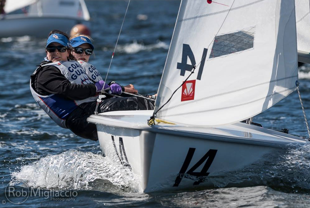 Two sailors wearing life vests and sunglasses lean out of a small sailboat marked with the number 14 as they compete on choppy water, with another boat visible in the background.